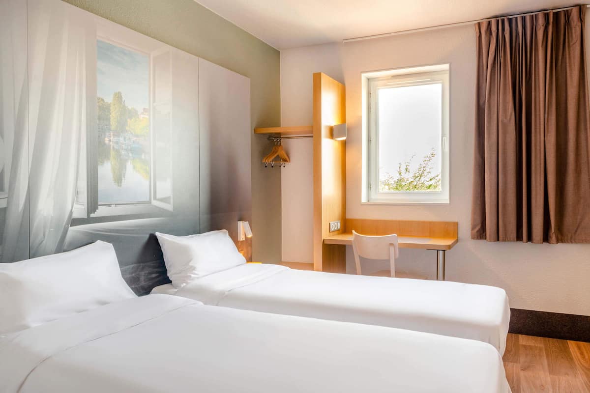 Ein Hotelzimmer mit zwei Betten, einem Schreibtisch, einem Stuhl, einem Fenster mit Vorhängen und einem Bild an der Wand.