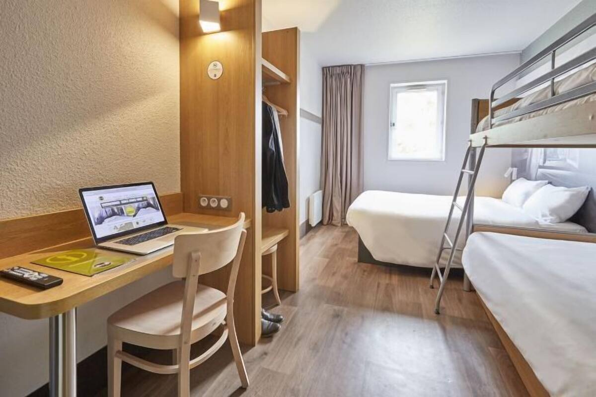 Ein kompaktes Hotelzimmer mit Etagenbett, Schreibtisch, Laptop, Stuhl und kleinem Kleiderschrank.