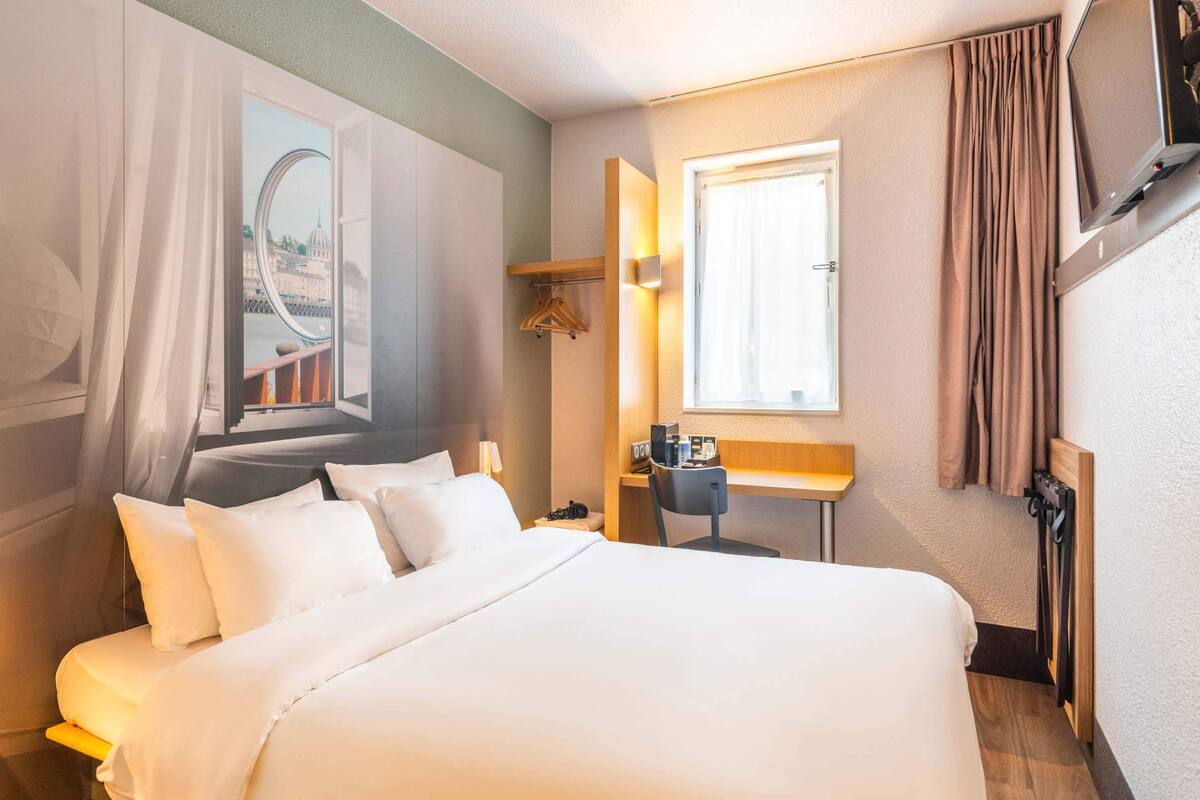 Ein Hotelzimmer mit einem großen Bett, einem Schreibtisch, einem Sessel, einem Fernseher und einem Fenster mit Vorhängen.