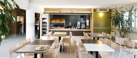 A modern cafe with multiple tables and chairs, a counter with food items, and a visible kitchen area.