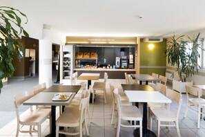 Ein modernes Café mit mehreren Tischen und Stühlen, einer Theke mit Speisen und einem sichtbaren Küchenbereich.