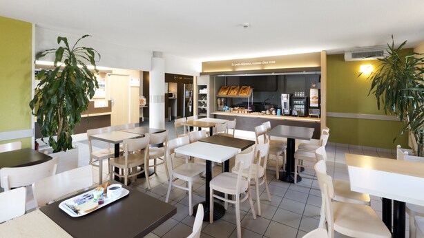 Un café moderne avec des tables et des chaises blanches, un comptoir où est affiché le menu, et des plantes en pot.