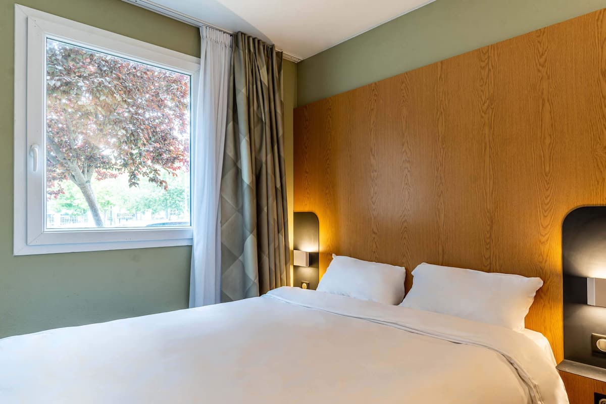 Een hotelkamer met een groot bed, een raam met gordijnen, een houten hoofdeinde en een bedlamp.