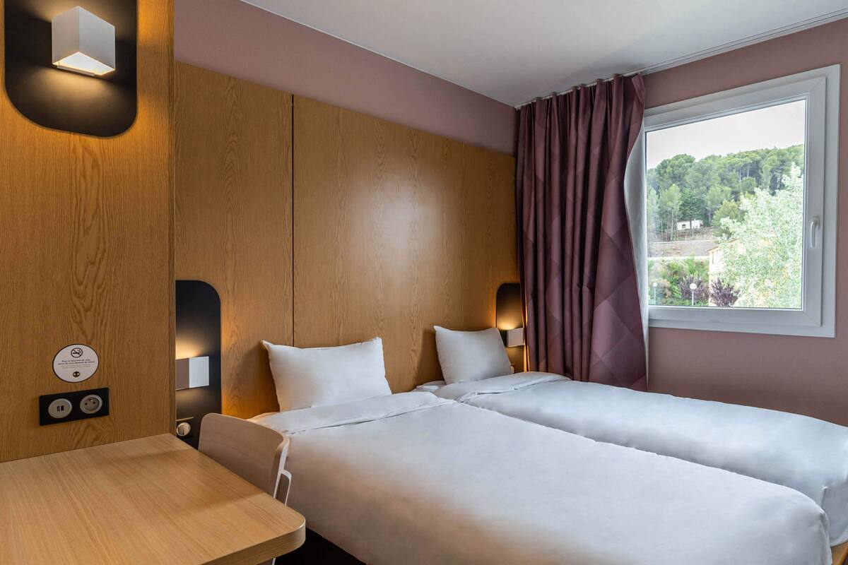 Hotelkamer met twee bedden, een houten hoofdbord, een raam met gordijnen en een tafel met stopcontact.