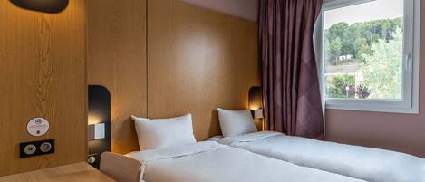 A hotel room with two beds, a wooden headboard, a window with curtains, and a table with a power outlet.