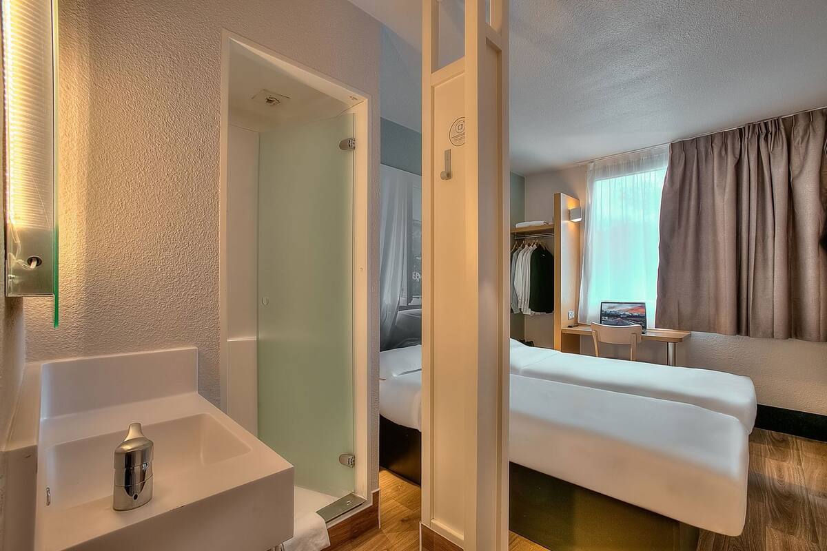 A hotel room with a shower, a bed, a bedside table, a wardrobe, and a window with curtains.