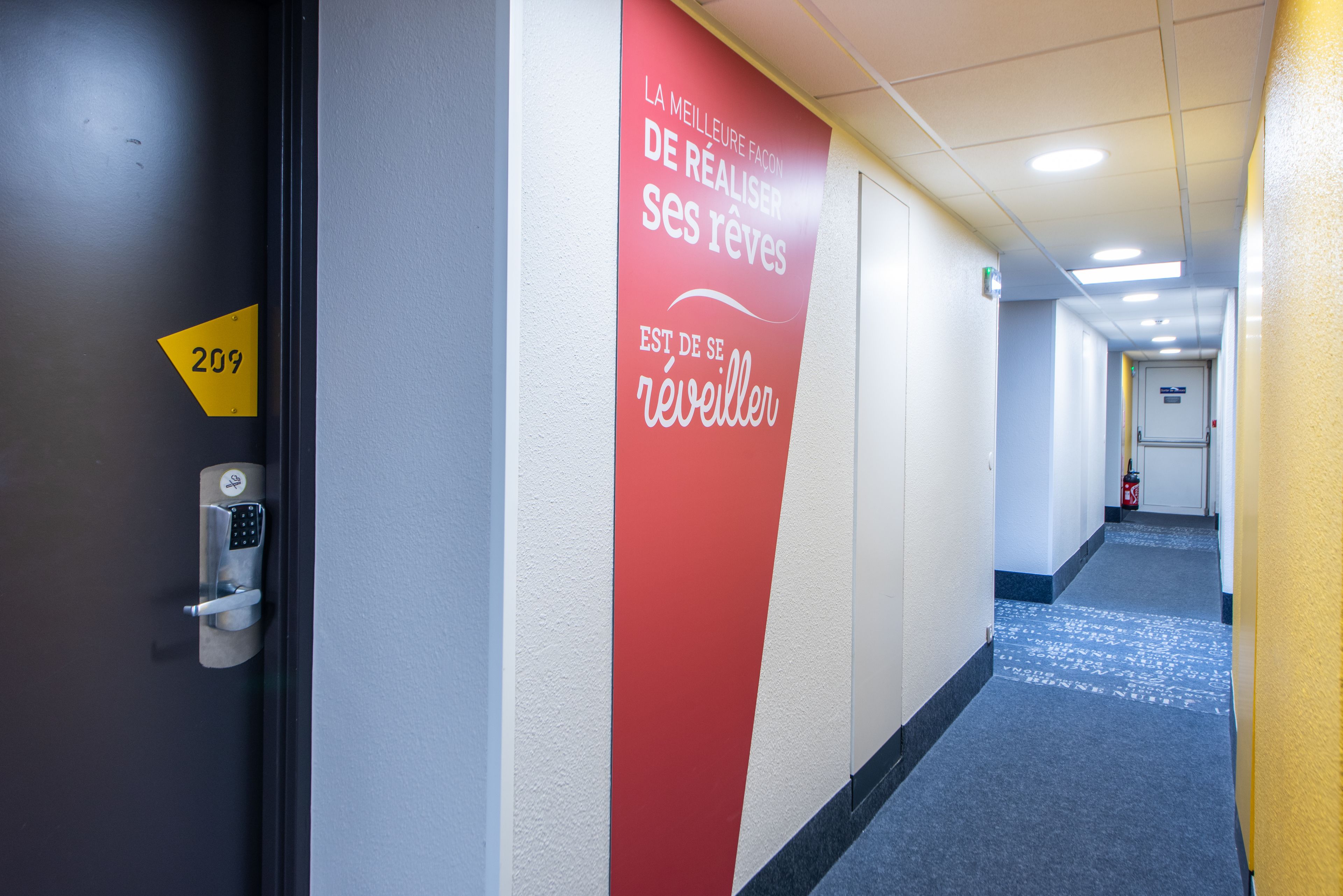 Un couloir d’hôtel avec un mur rouge orné d’un texte motivant, une porte portant un numéro et un tapis bleu.