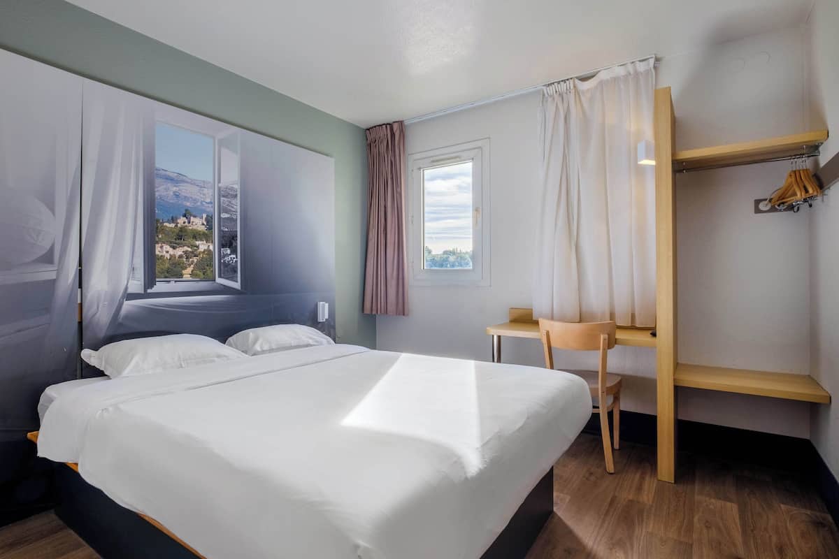 A hotel room with a bed, a desk, a chair, and a view of a mountainous landscape.