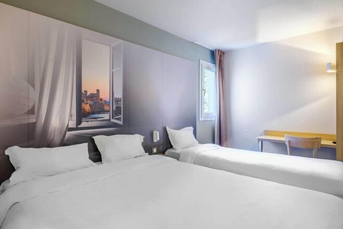 A hotel room with two beds, a desk, a chair, and a large window with a cityscape view.