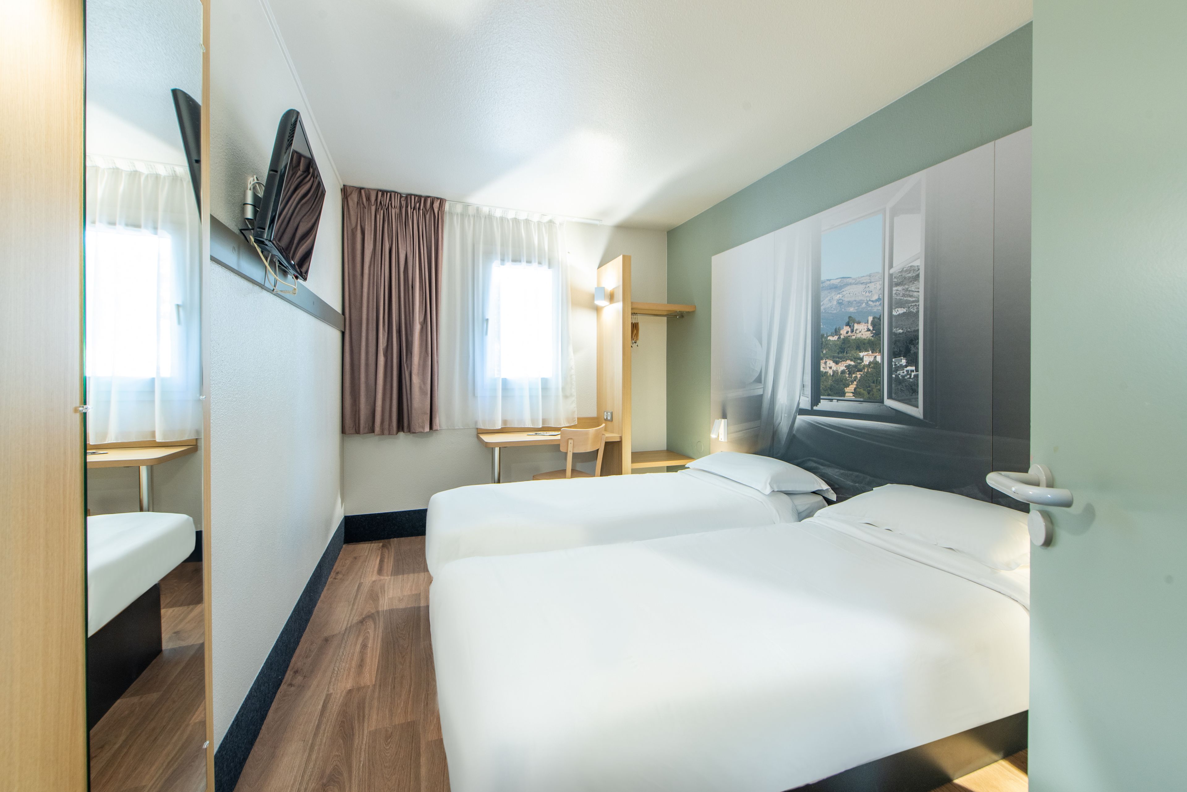A hotel room with two beds, a desk, a chair, and a window with a view.