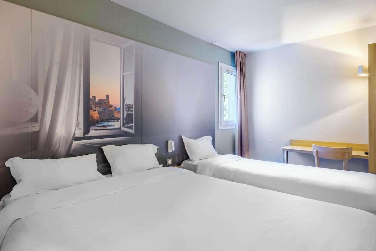 A hotel room with two beds, a desk, and a view of a cityscape through a window.