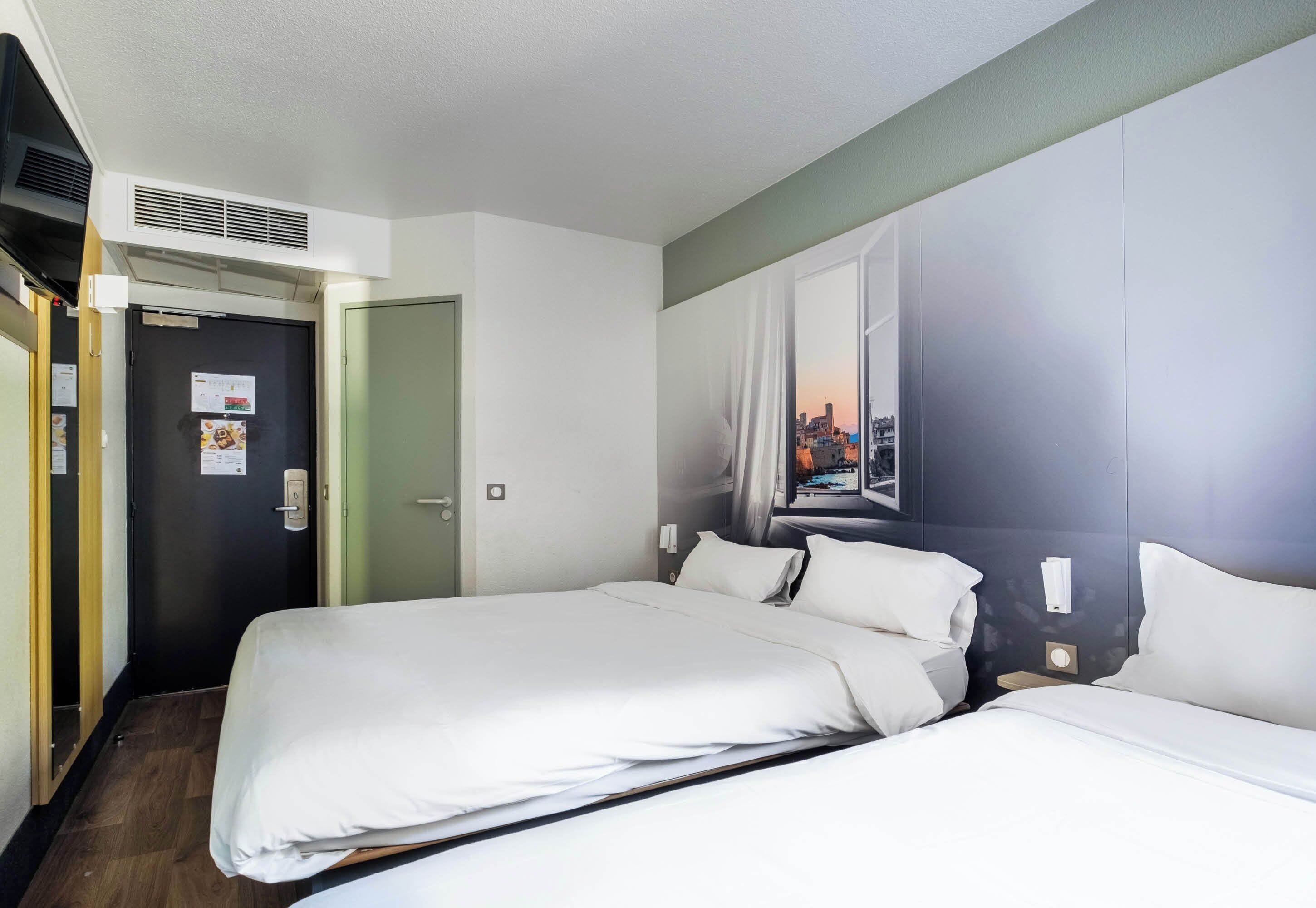 Une chambre d’hôtel avec deux lits, une grande fenêtre donnant sur la ville, une porte ornée d’un calendrier et un climatiseur fixé au mur.
