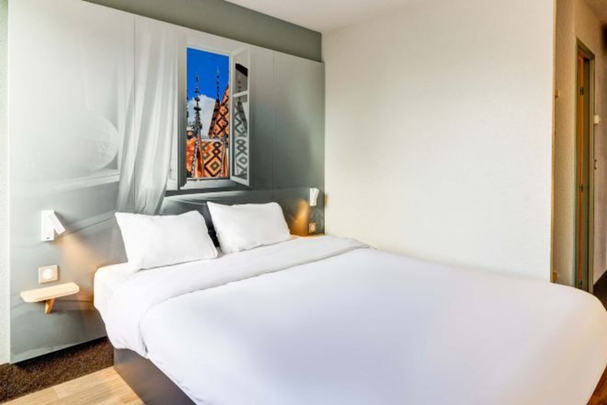 A modern hotel room with a large bed, a nightstand, a wall-mounted lamp, and a window with a view of a building.
