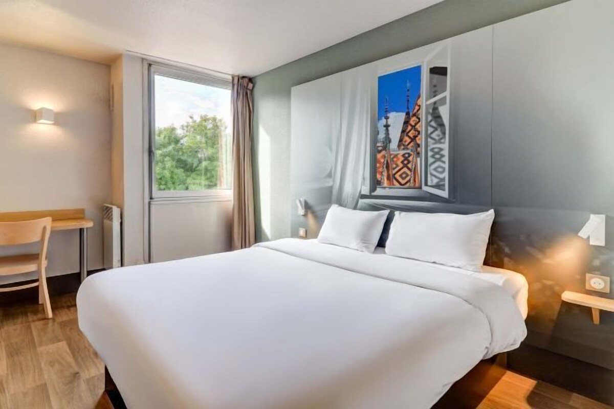 A hotel room with a large bed, a desk, and a window with a view of greenery.