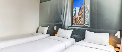 Habitació d'hotel amb dos llits, una finestra gran amb vista a un edifici i una obra d'art penjada a la paret.
