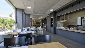 A modern restaurant with a mix of indoor and outdoor seating, a kitchen area, and large windows offering views of the outdoors. - B&B HOTEL Beaune Sud (1) Palais des Congrès (Beaune)