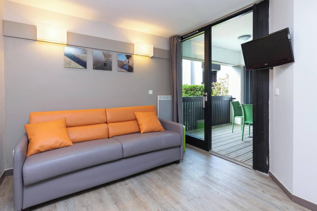 Una habitación con sofá gris, televisor de pantalla plana y puerta de vidrio corredera que da a un espacio exterior con una silla verde.