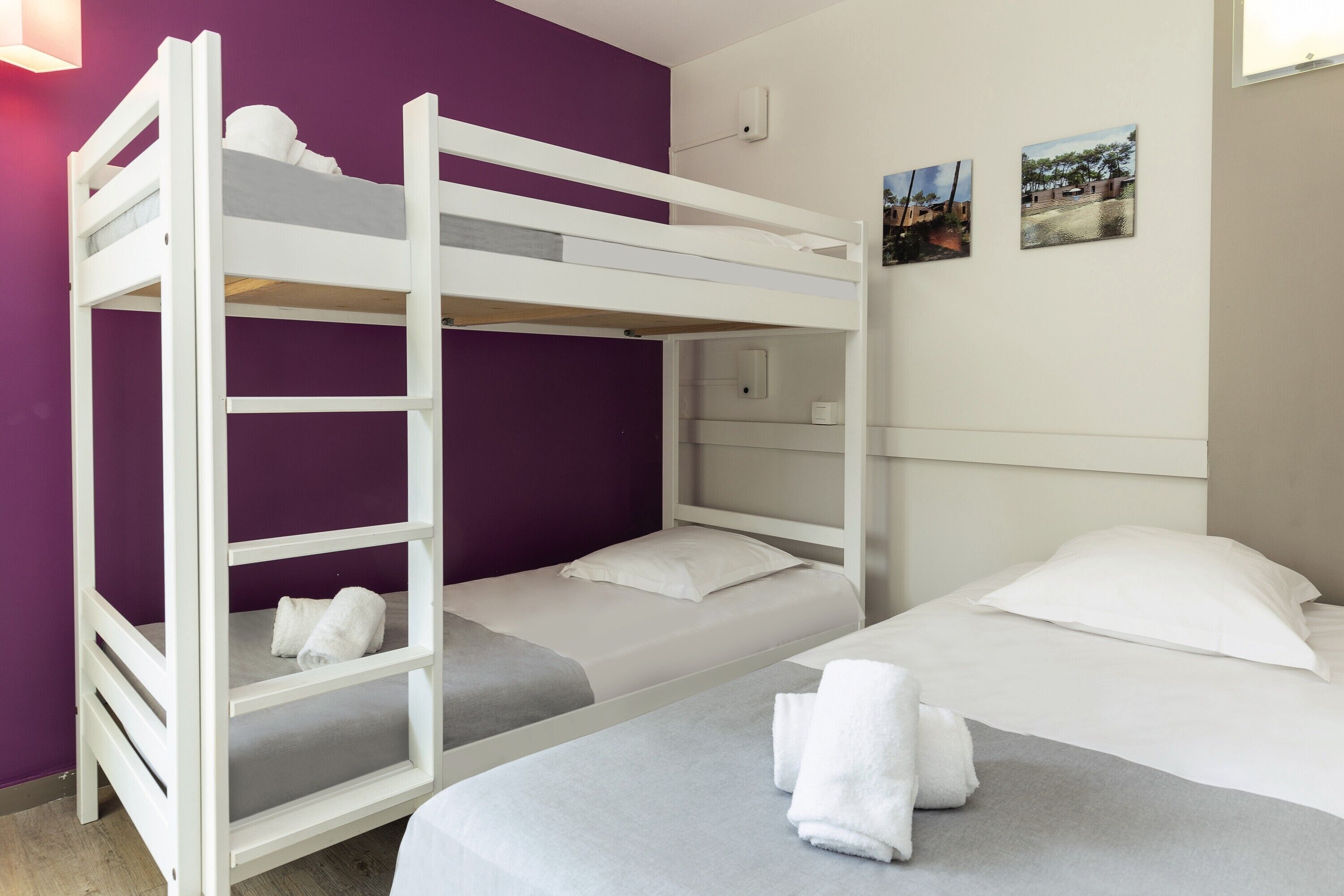 Une chambre avec un lit superposé, un mur violet, un lit blanc et deux petits tableaux accrochés au mur.