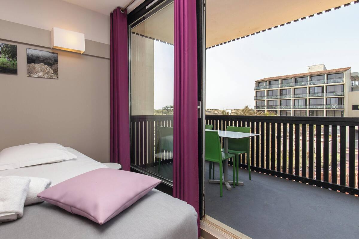 Habitación de hotel con cama, ventana con cortinas, mesa con sillas y vista del edificio exterior.
