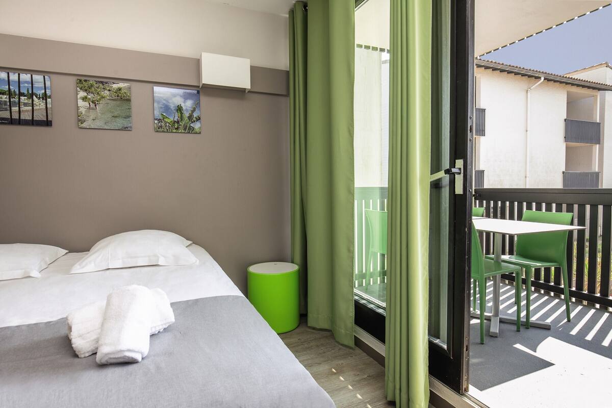Una habitación de hotel con una cama, un cubo de basura verde y un balcón con una mesa y sillas.