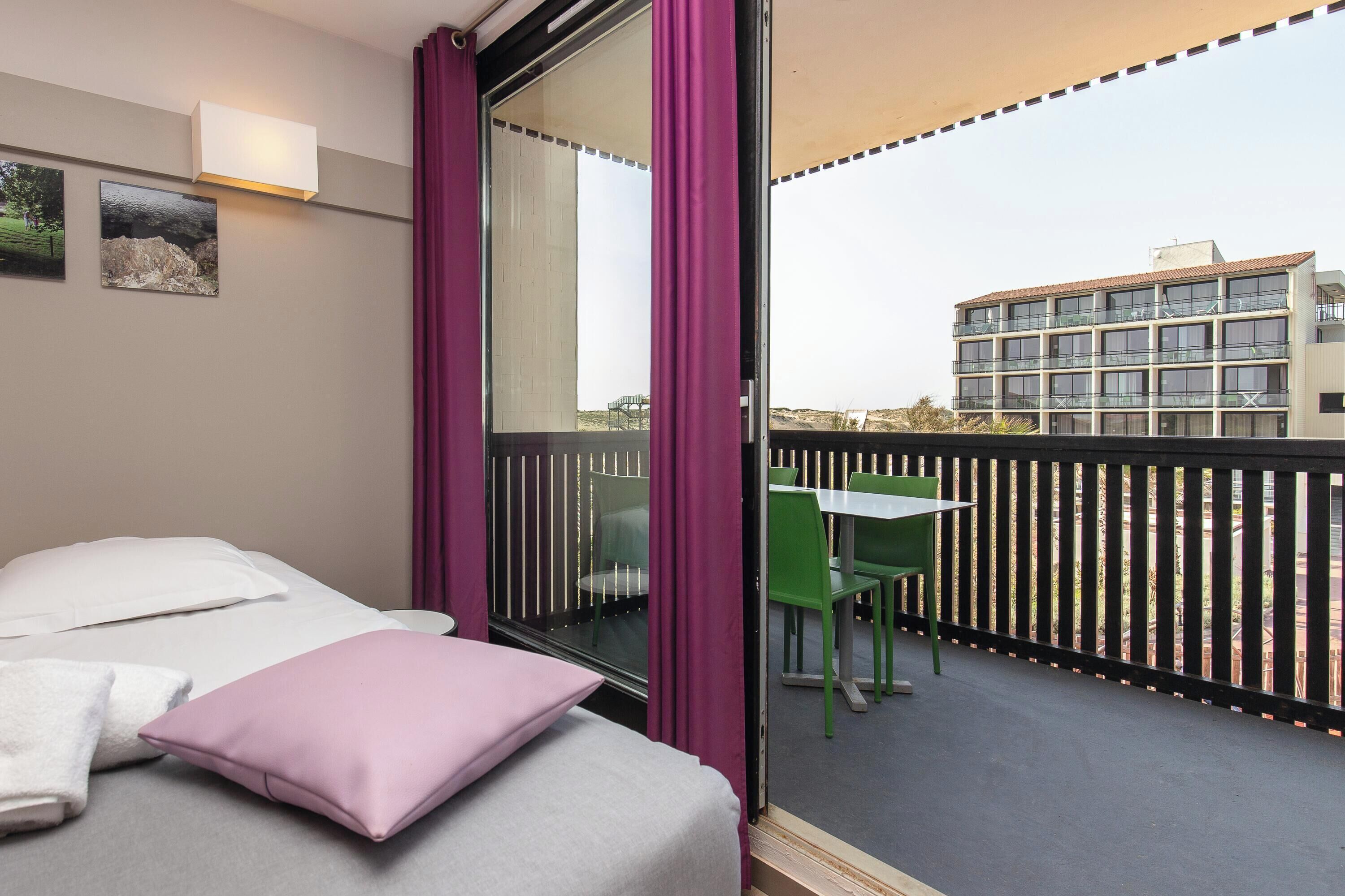 A hotel room with a bed, a window with curtains, a table with chairs, and a view of the building outside.