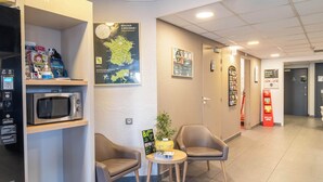 A hotel reception area with a reception desk, a microwave, two chairs, a small table with a plant, and various informational posters on the walls. - B&B HOTEL Brive-la-Gaillarde (Ussac)