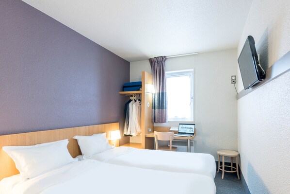 Habitación de hotel con dos camas, un escritorio con una computadora portátil, un televisor montado en la pared y una ventana con cortinas.