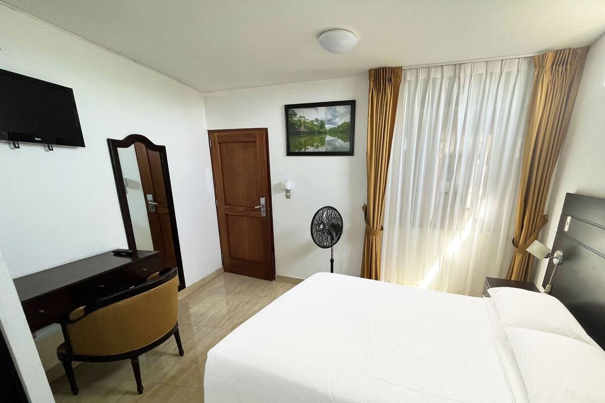A hotel room with a bed, a desk, a chair, a mirror, a door, a fan, and a window with curtains.