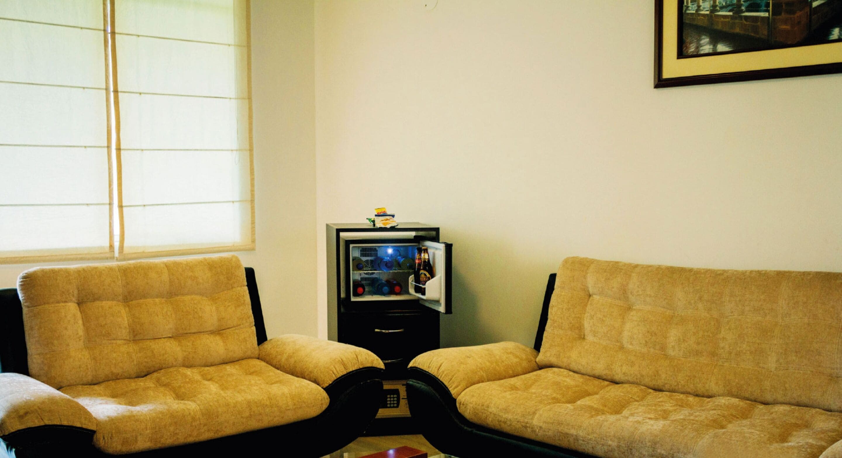 Een kamer met twee beige banken, een kleine koelkast en een raam met jaloezieën.
