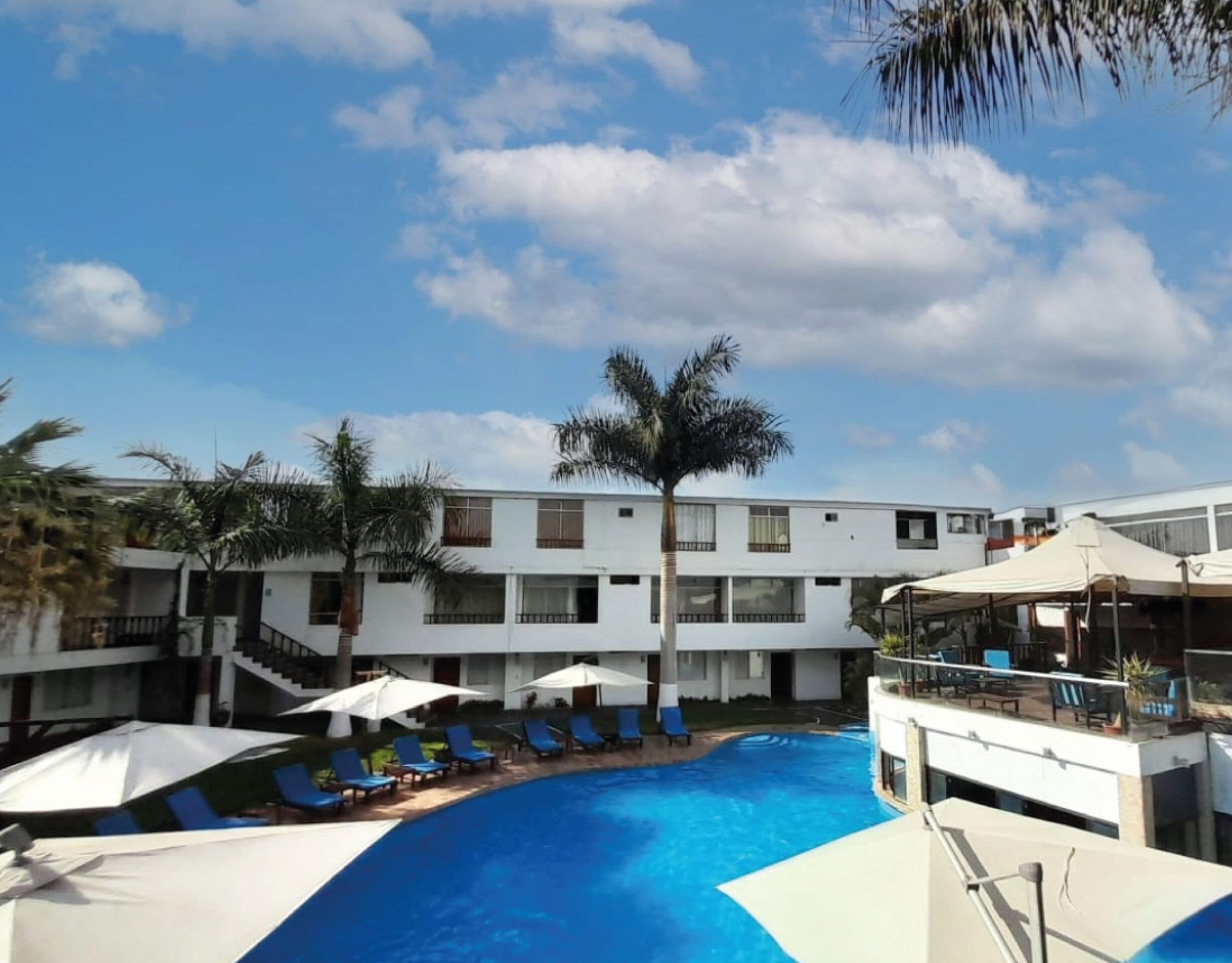 A pool area with blue water, white umbrellas, and blue lounge chairs. There are palm trees and a building with multiple windows and balconies in the background.