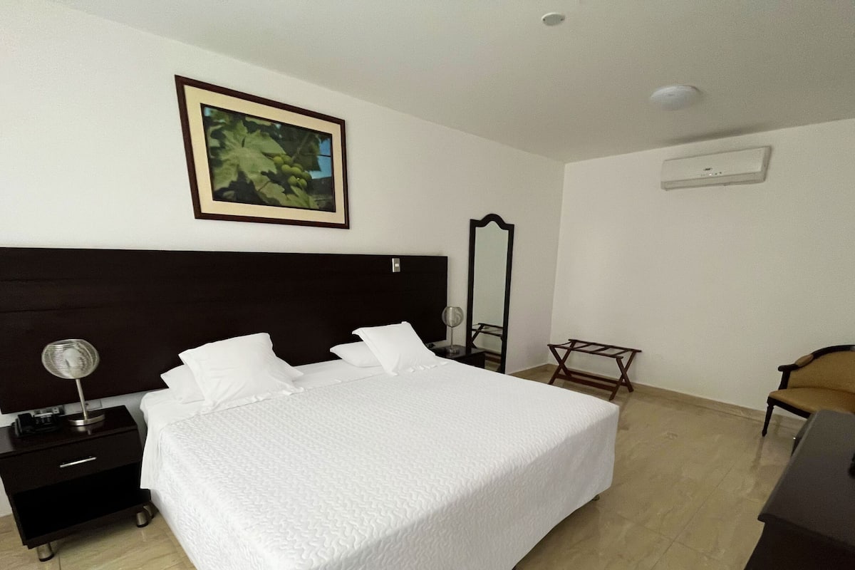 A hotel room with a large bed, a nightstand, a chair, a desk, and a framed picture on the wall.