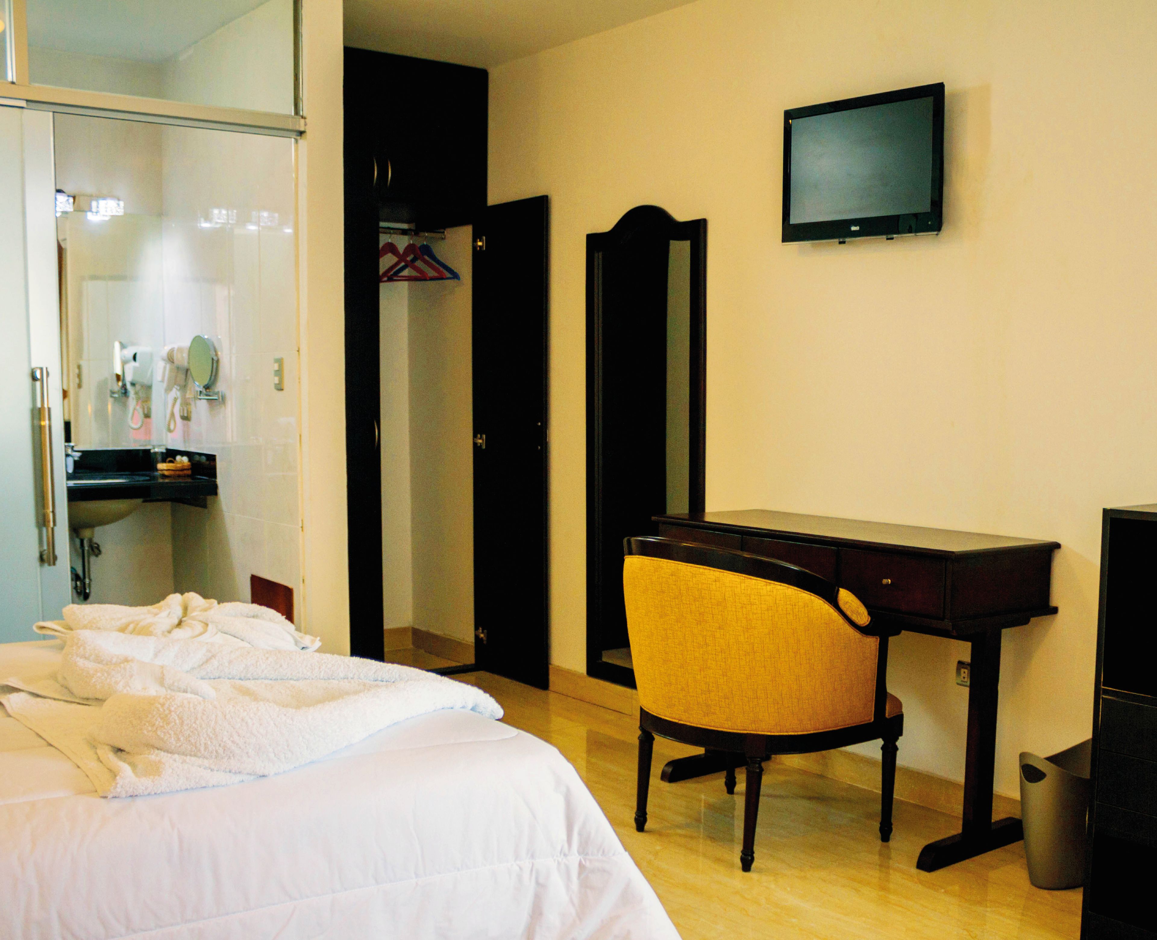 Quarto de hotel com cama, escrivaninha, cadeira, televisão e guarda-roupa.