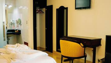 Quarto de hotel com cama, escrivaninha, cadeira, televisão e guarda-roupa.