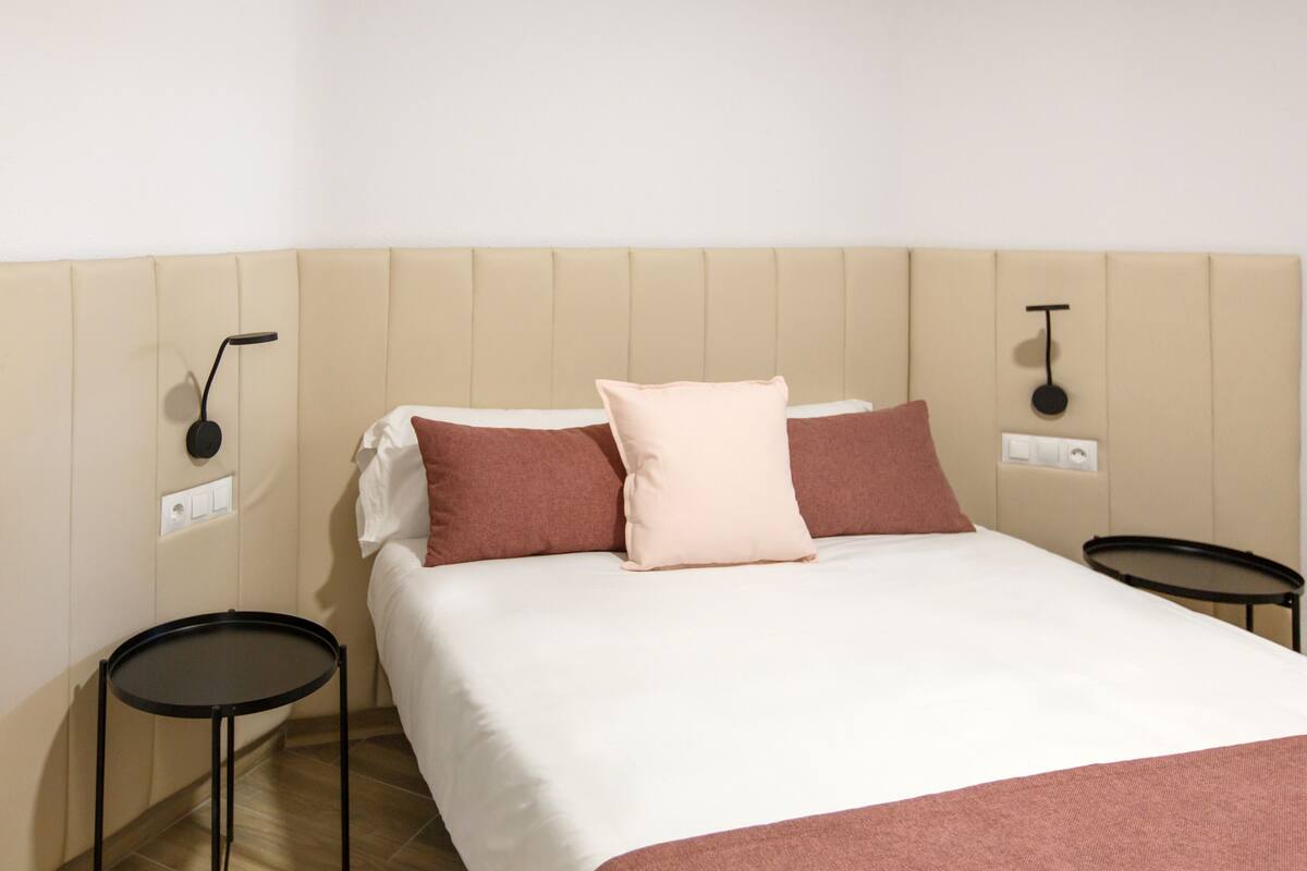 A neatly made bed with a white and pink pillow arrangement, two bedside tables, and a wall-mounted light fixture.