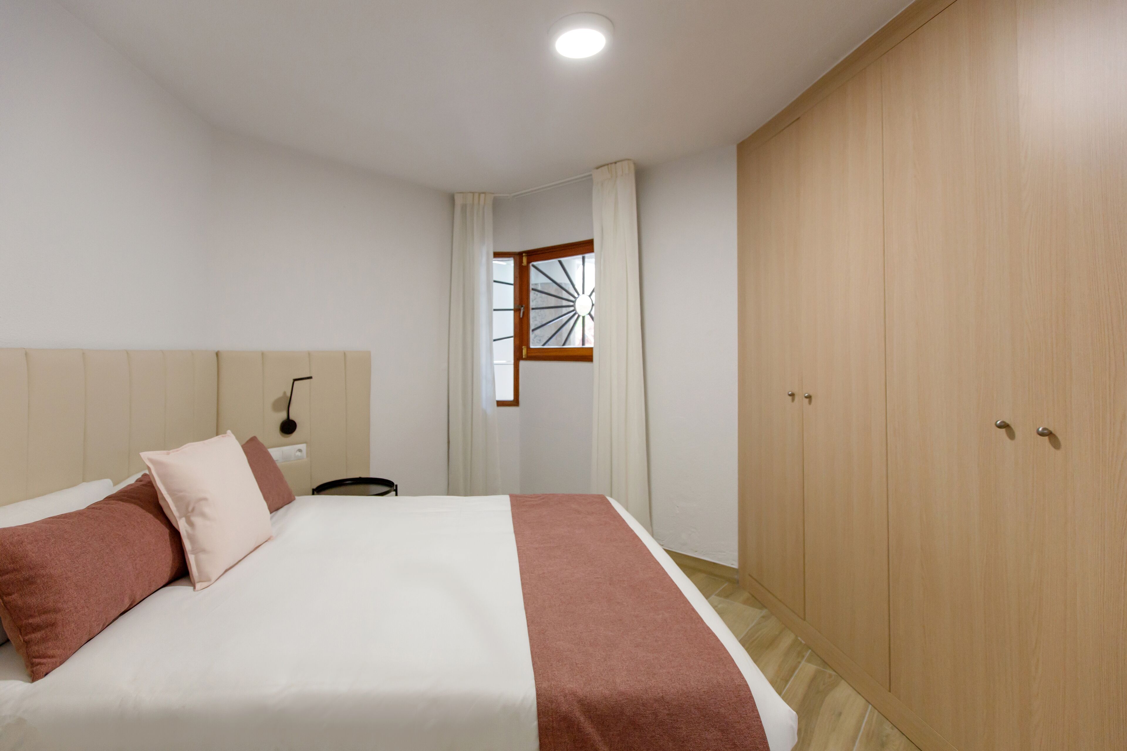 A bedroom with a bed, a wardrobe, a window with curtains, and a wall-mounted light fixture.