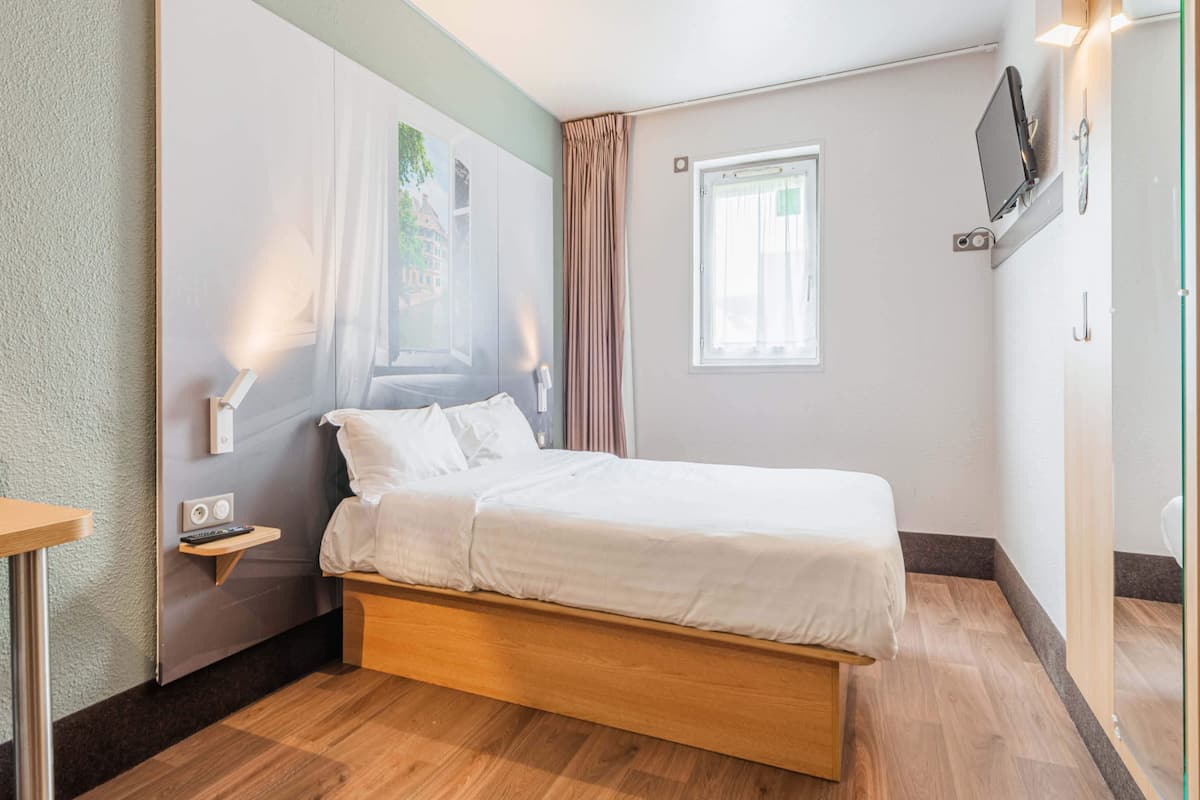 Un dormitorio ordenado con una cama individual, una mesita de noche, un televisor montado en la pared y una ventana con cortinas.