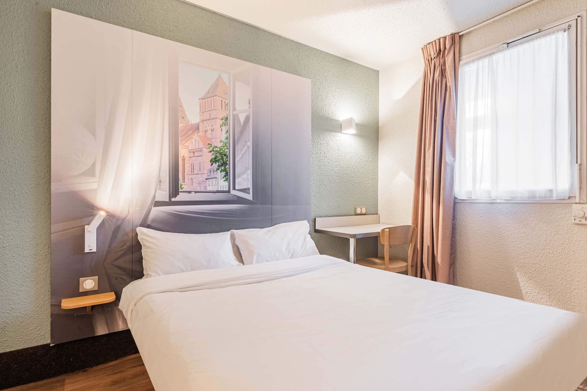 Una habitación de hotel con cama, una mesita pequeña, una silla, una mesita de noche y una lámpara fijada en la pared.