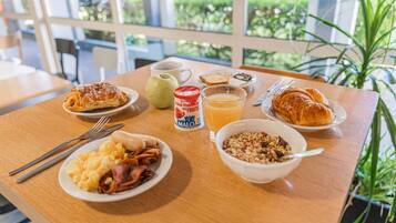 A breakfast table with croissants, scrambled eggs, ham, a bowl of cereal, a cup of coffee, a glass of orange juice, and a can of fruit yogurt.