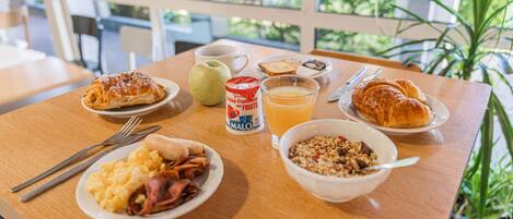 Una mesa de desayuno con cruasanes, huevos revueltos, jamón, un tazón de cereal, una taza de café, un vaso de jugo de naranja y una lata de yogur de frutas.