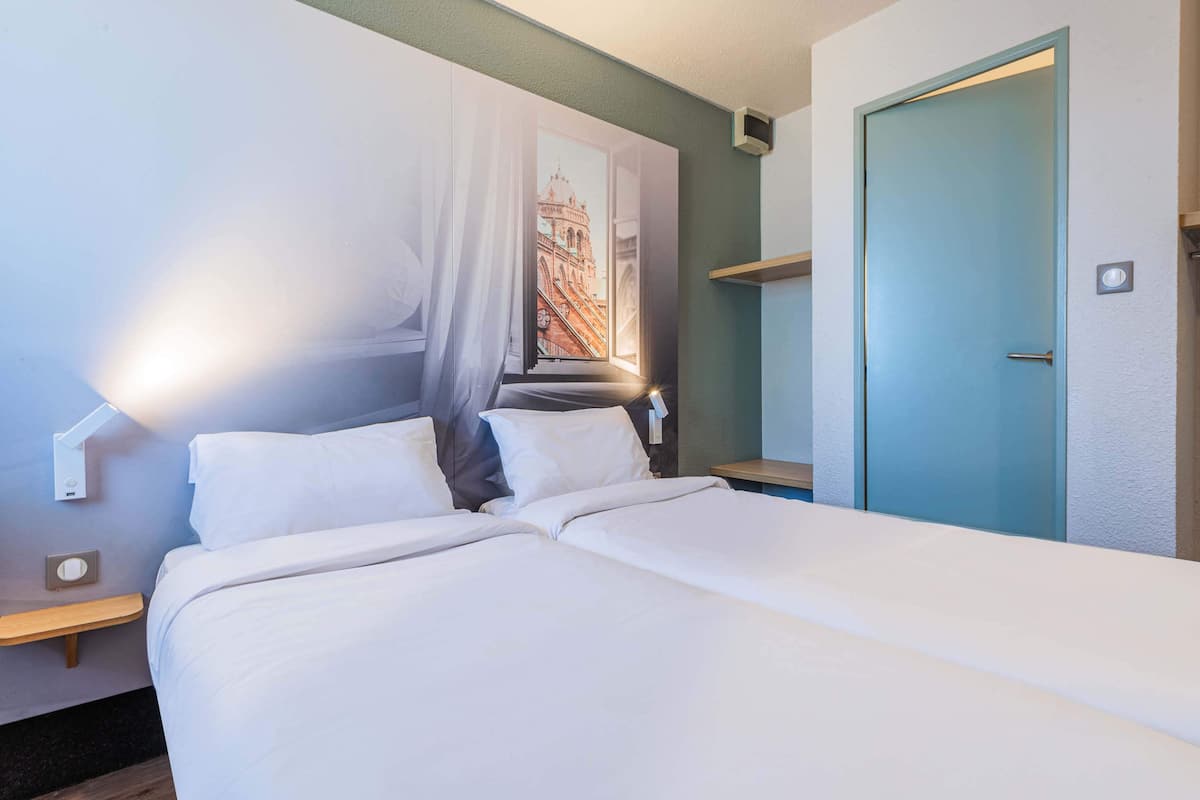 Habitación de hotel con dos camas, una lámpara de pared, una puerta y una ventana con vista a un edificio.