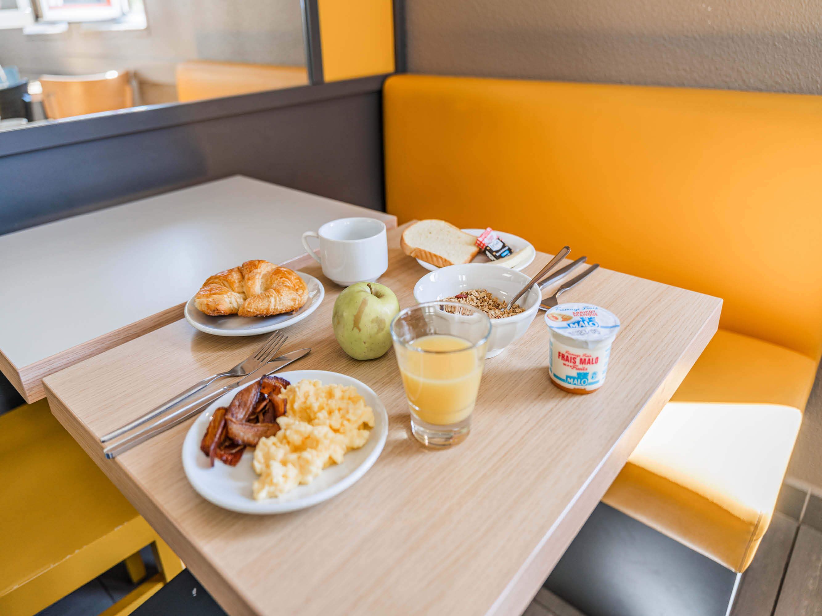 Una mesa de desayuno con cruasanes, huevos revueltos, tocino, un tazón de cereal, un vaso de jugo de naranja y un recipiente de yogur.