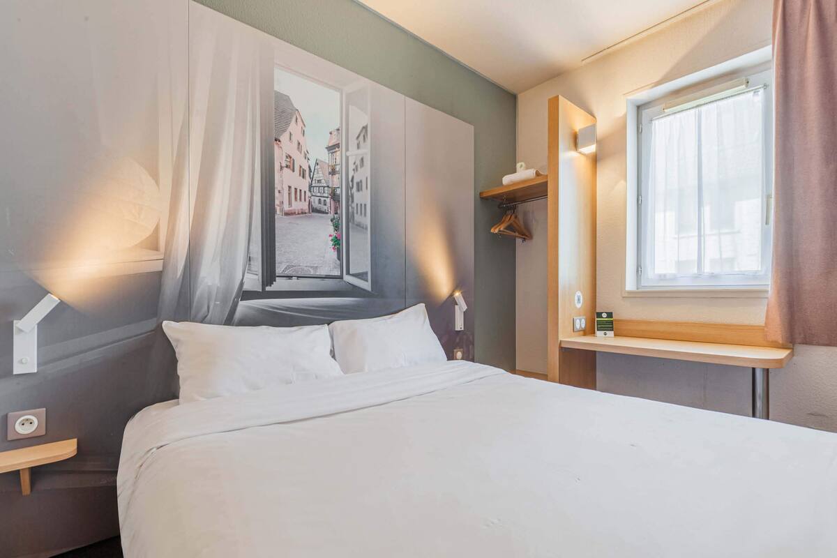 A neatly made bed in a hotel room with a view of a street through a window, a bedside table with a lamp, and a small shelf with a towel.