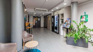 A modern lobby with a seating area, a vending machine, a green emergency exit sign, and a potted plant. - B&B HOTEL Strasbourg Sud Ostwald (Ostwald)