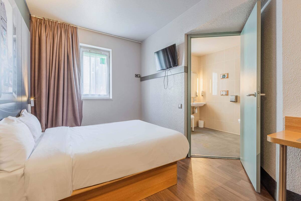 A bedroom with a bed, a TV mounted above it, and a bathroom visible through an open door.