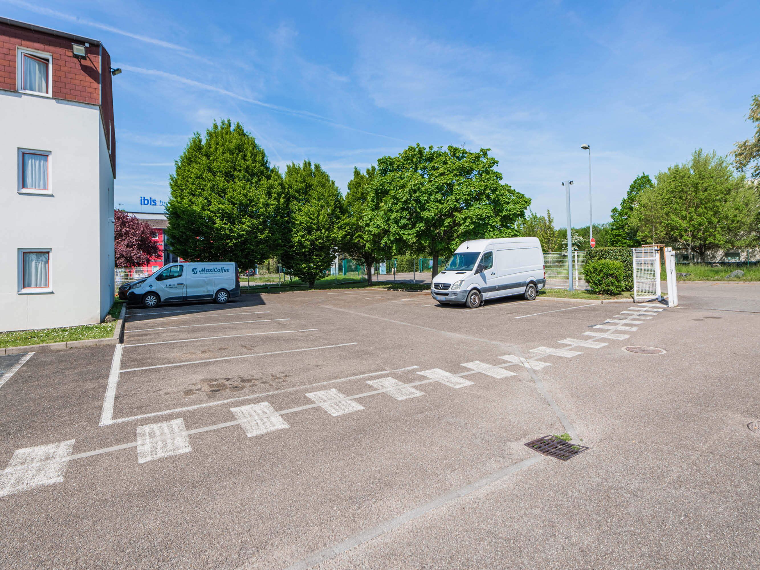 Een parkeerplaats met twee witte bestelwagens, waarvan er een 'MaxiCoffee' op de bestelwagen heeft staan, en een gebouw met 'ibis' aanduiding.
