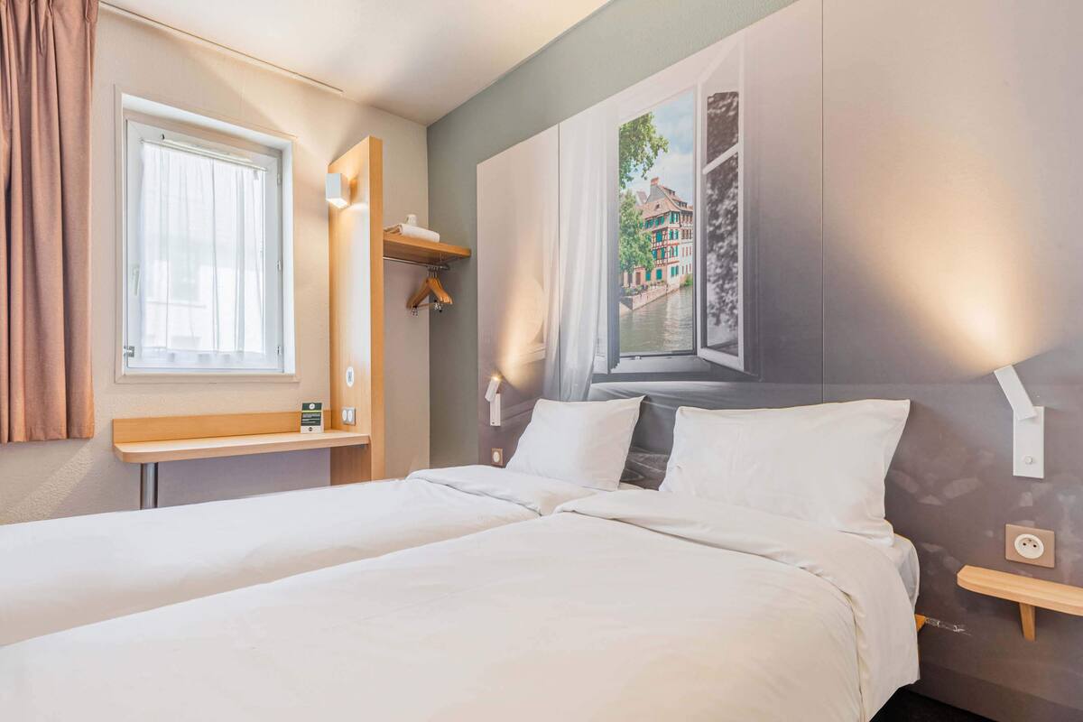 A hotel room with two beds, a window with a view of buildings, a wooden shelf, and a wall-mounted light.