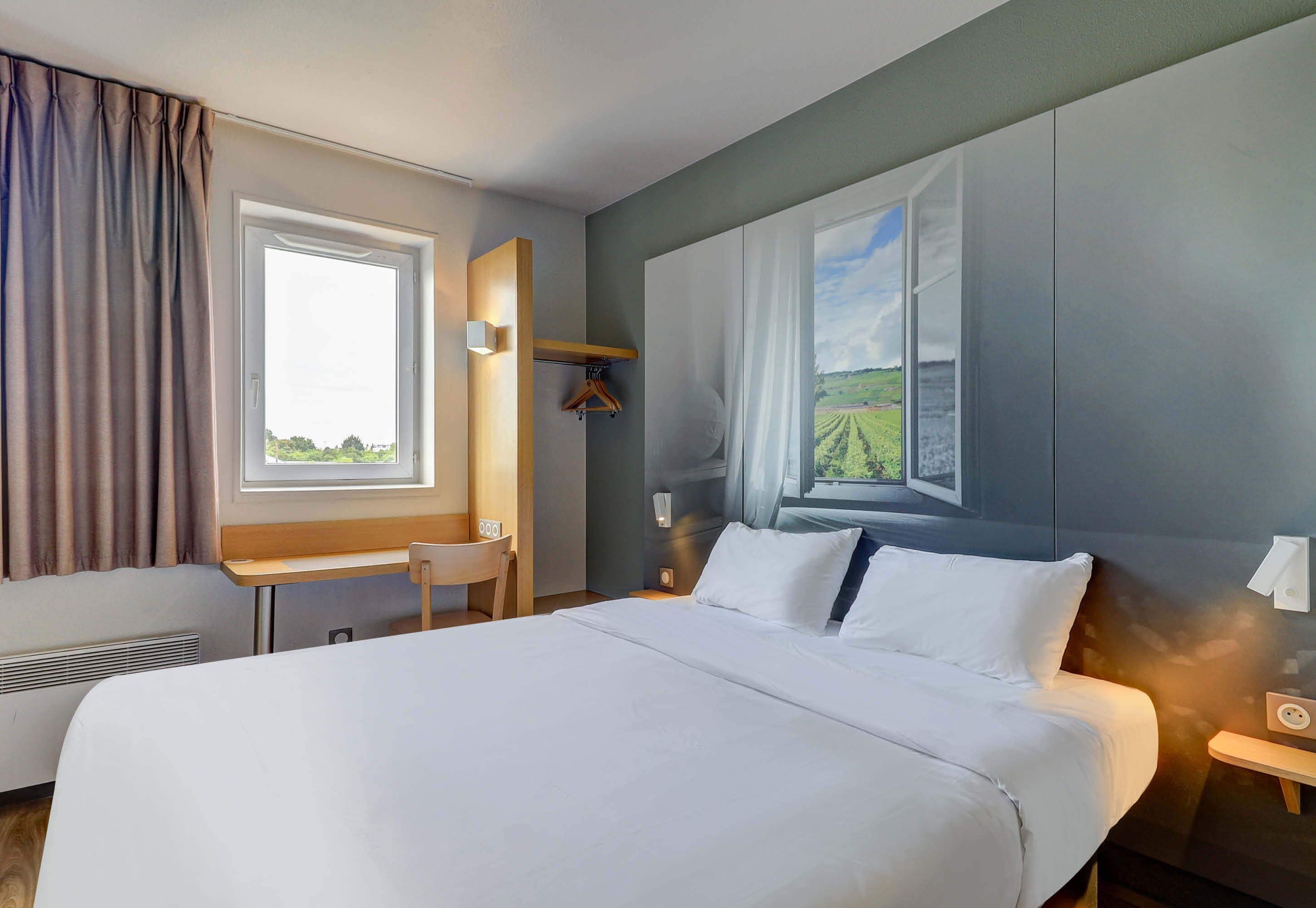 Une chambre d’hôtel avec un grand lit, un bureau et une chaise. Il y a une fenêtre donnant sur un champ.