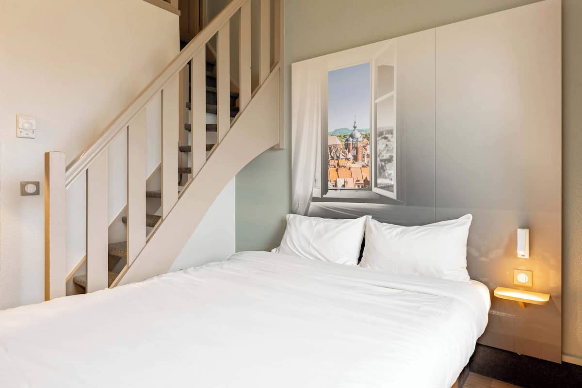 A hotel room with a bed, a window with a cityscape view, a staircase, and a wall-mounted light fixture.