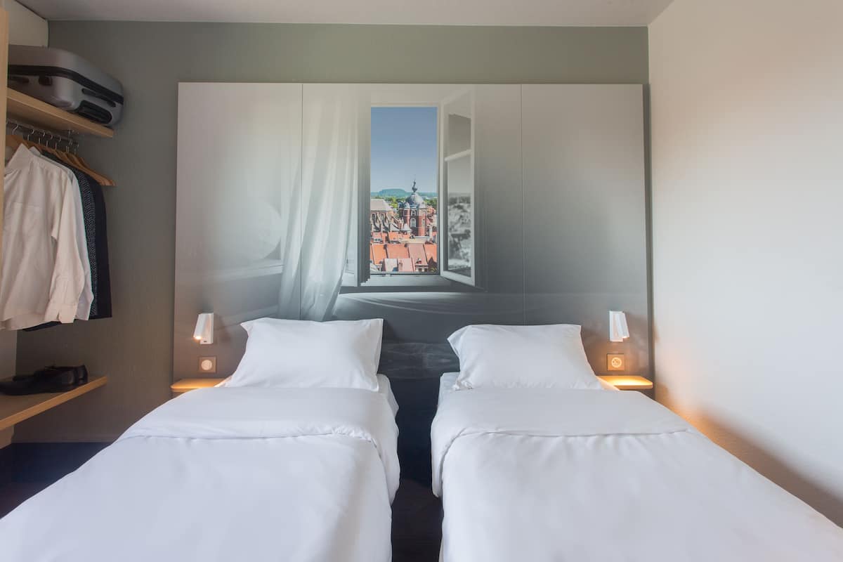 A hotel room with two beds, a wardrobe, a shelf with a coat, and a painting of a cityscape.