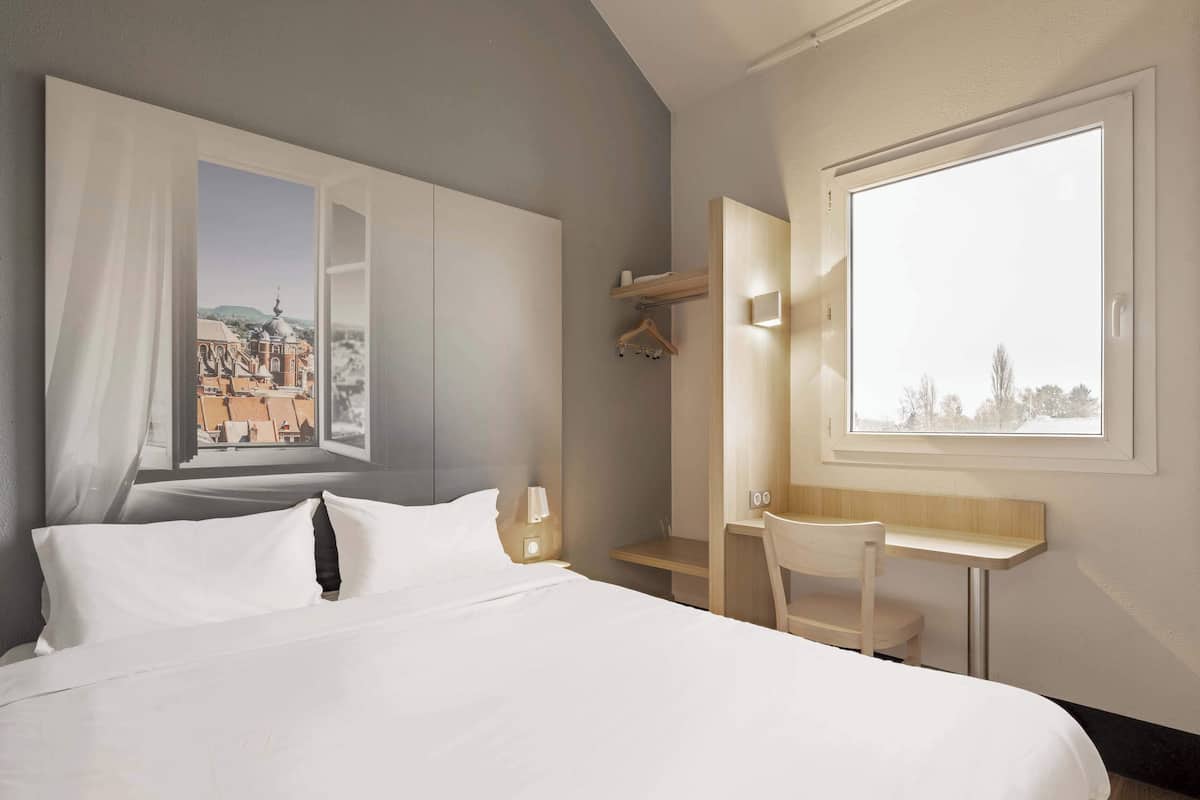 A hotel room with a large bed, a desk, a chair, a wardrobe, and a window with a view of buildings.